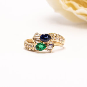 Sormuksen keskiössä loistavat ovaali viistehiottu sininen safiiri ja viistehiottu vihreä smaragdi, jotka asettuvat kauniisti toisiaan täydentäviksi.